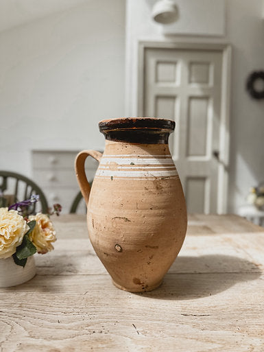 A vintage Hungarian jug made from terracotta with white stripes around the rim. The jug is set on a rustic wooden table next to a small pot of flowers.