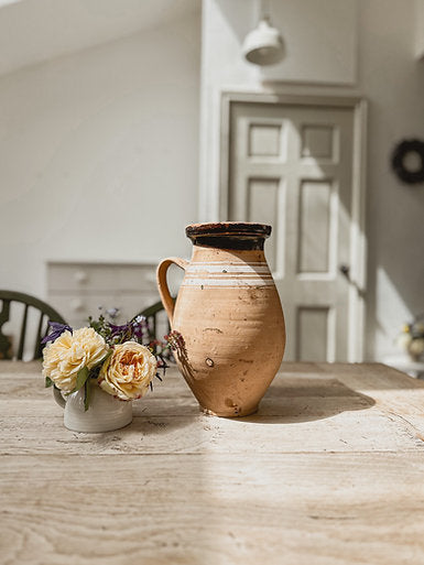 An antique hungarian jug is sat atop a rustic wooden table in a large and bright airy room. The room is painted white and the woodwork and door are painted in Farrow & Ball Shaded White.