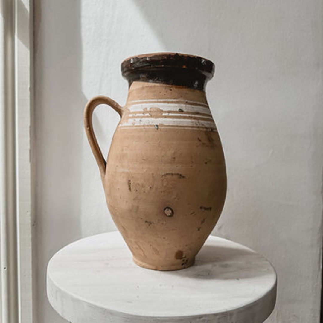 Vintage Hungarian terracotta pot with handle. The jug is placed on top of a whitewashed stool and set against a white wall.